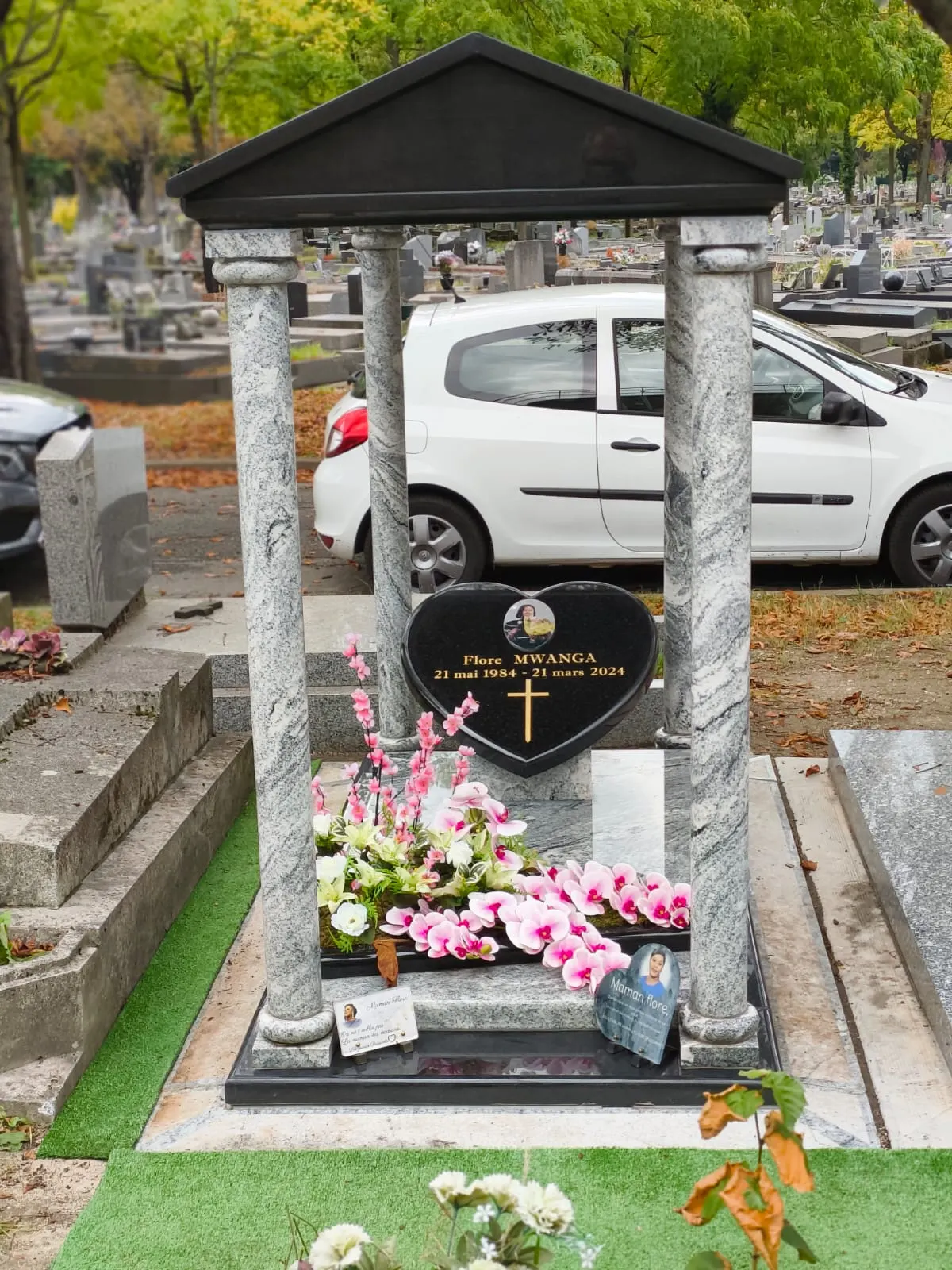Vue d’un monument avec deux colonnes, un fronton sombre, une plaque en cœur et des compositions florales.